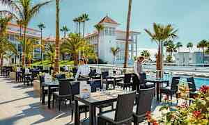 Restaurantområde med udendørs siddepladser, omgivet af palmer og swimmingpools. (AI-genereret tekst)