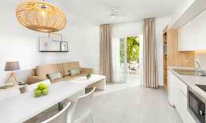 Et lyst og moderne møbleret rum med en beige sofa, hvidt spisebord og køkken samt åbne døre til en møbleret altan. (AI-genereret tekst)