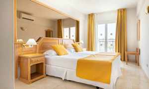 Et hotelværelse med en dobbeltseng, hvide lagner og gule puder og sengetæppe. Værelset har lyse trémøbler og vinduer med gardiner. (AI-genereret tekst)