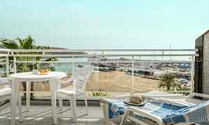 Udsigt fra en altan med hvide havemøbler, med blik over en strand, en lystbådehavn og havet i baggrunden. (AI-genereret tekst)