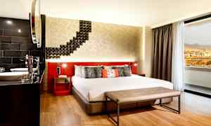 Et hotelværelse med en stor seng, moderne indretning og en siddebænk foran sengen. Et vindue viser udsigten til bjerge og bygninger. (AI-genereret tekst)