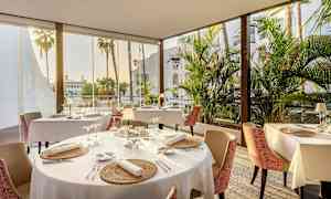 En restaurant med dækkede borde, store vinduer og udsigt over palmer. (AI-genereret tekst)