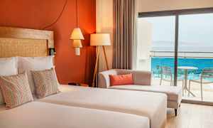 Et hotelværelse med dobbeltseng, dekorative puder, en sofa og glasdøre, der fører til balkonen med udsigt over havet. (AI-genereret tekst)