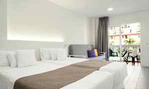 Et hotelværelse med to enkeltsenge med hvidt sengetøj, en grå sofa med farvede puder og en balkon med udsigt. (AI-genereret tekst)