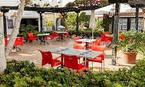 Udendørs serveringsområde med røde stole og borde, omkranset af beplantning. (AI-genereret tekst)