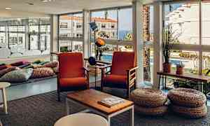 Et lyst rum med store vinduesdøre, to orange lænestole og kurvskamler foran et kaffebord. (AI-genereret tekst)