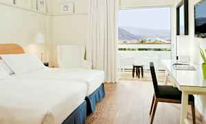Et lyst og moderne hotelværelse med to enkeltsenge, en hvid lænestol og et skrivebord med lampe. Et stort vindue fører ud til en balkon med udsigt over bjerge og grønt. (AI-genereret tekst)