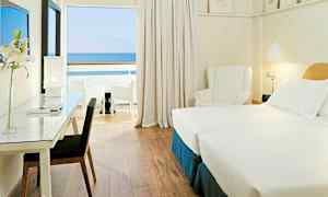 Et hotelværelse med to enkeltsenge, skrivebord med stol, en hvid lænestol og udsigt til havet fra en privat altan. (AI-genereret tekst)