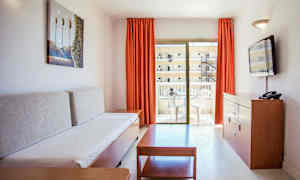 Stue i en ferielejlighed med en sofa, et sofabord, en kommode med et TV og en altan med havemøbler. (AI-genereret tekst)