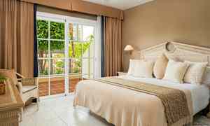 Et hotelværelse med en stor seng, beige sengetøj, hvide puder og en glasdør, der åbner ud til en terrasse med beplantning. (AI-genereret tekst)