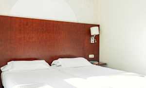 To enkeltsenge med hvidt sengetøj og en mørk træhovedgavl. En væglampe er monteret på siden. (AI-genereret tekst)