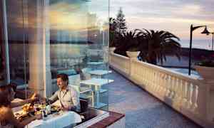 Par, der spiser middag ved et bord nær glasvæg med udsigt over terrasse og hav. (AI-genereret tekst)