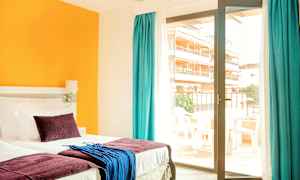 Et hotelværelse med en seng, farverige puder og tekstiler samt en glasdør, der fører til en altan. (AI-genereret tekst)