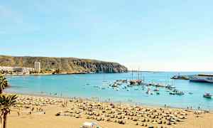 Strand med liggestole og parasoller ved en beskyttet havbugt med både og bygninger synlige i baggrunden. (AI-genereret tekst)