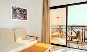 Udsigt fra et hotelværelse med en beige sofa, et træbord og en altan med udsigt over stranden. (AI-genereret tekst)