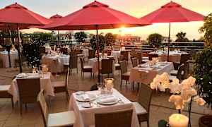 Udendørsrestaurant med røde parasoller, dækkede borde og solnedgang i baggrunden. (AI-genereret tekst)