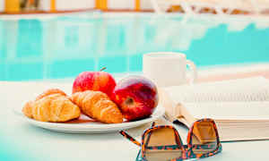 En tallerken med croissanter og æbler står på et bord ved siden af en bog og et par solbriller. I baggrunden er en pool synlig. (AI-genereret tekst)