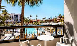 Udsigt fra en altan over et hotelområde med pool og liggestole, omgivet af palmer. (AI-genereret tekst)