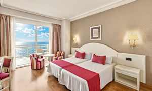 Et hotelværelse med to senge, røde accenter og havudsigt fra en altan. (AI-genereret tekst)