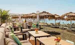 Udendørs terrasse med siddepladser og borde med udsigt til en strand med parasoller og liggestole. (AI-genereret tekst)