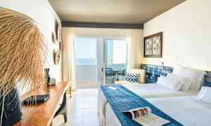 Et lyst hotelværelse med to senge, en træbænk og en altan med havudsigt. (AI-genereret tekst)