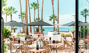 Restaurantmiljø med udendørs servering, møbler og palmer med havudsigt. (AI-genereret tekst)