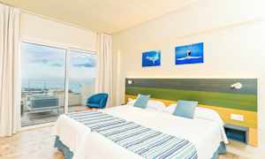 Hotelværelse med to enkeltsenge, en grøn og blå sengegavl samt en balkon med udsigt over havet. (AI-genereret tekst)