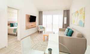 Billedet viser en lys og moderne stue med en sofa, fjernsyn samt spisebord og stole. En dør fører til et soveværelse. (AI-genereret tekst)