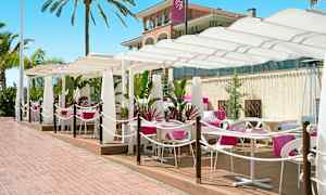 Udendørs servering med hvide parasoller og lyserøde duge på bordene. Palmer og en bygning i baggrunden. (AI-genereret tekst)