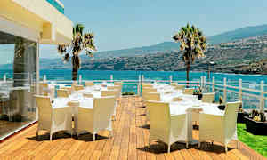 Udendørs terrasse med spiseborde og stole, udsigt over havet og det omkringliggende landskab. (AI-genereret tekst)