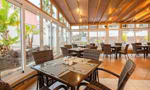 En spisesal med kurvestole og træborde, omgivet af store glasvinduer med udsigt til en have. (AI-genereret tekst)