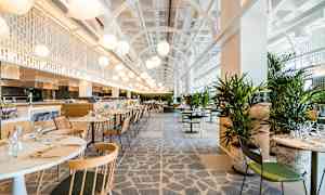 En moderne og lys restaurant med borde og stole samt grønne planter. Loftshøjde og belysning bidrager til en luftig atmosfære. (AI-genereret tekst)