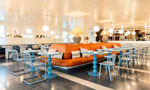 En moderne indrettet café med orange lædersofaer, blå borde og stole samt dekorative lamper i loftet. (AI-genereret tekst)