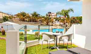 Udsigt fra en terrasse med liggestole, bord og stole mod en pool og omkringliggende grøntområder. (AI-genereret tekst)