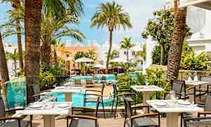 Billede af en restaurantterrasse med udsigt til en pool omgivet af palmer og lyse bygninger. (AI-genereret tekst)