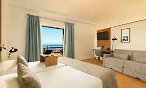 Et hotelværelse med en seng, sofa, skrivebord og et stort vindue med udsigt over havet. (AI-genereret tekst)