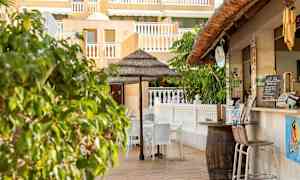 Udendørs serveringsområde med stole og en parasol nær en bygning med gul facade og hvide rækværk. (AI-genereret tekst)