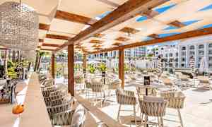 En udendørsbar med stole og borde under en pergola, omgivet af liggestole og grønt. (AI-genereret tekst)