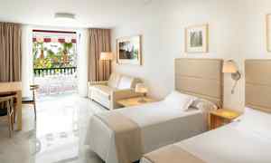 Hotelværelse med to enkeltsenge, en sofa, et bord med stole og et vindue mod en altan. (AI-genereret tekst)