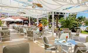 Udendørs restaurant med solafskærmning, borde og stole samt grønt i baggrunden. (AI-genereret tekst)