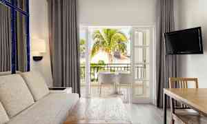 Et opholdsområde med stor sofa, træbord og en TV, der åbner ud mod en balkon med udsigt over tropiske træer og bygninger. (AI-genereret tekst)