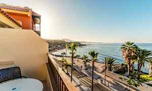 Udsigt fra en balkon mod en palmeomkranset strandpromenade og havet under en klar himmel. (AI-genereret tekst)