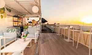 En solrig tagterrasse med stole, bar og borde samt solnedgang i baggrunden. (AI-genereret tekst)