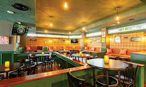 Et hyggeligt indrettet café eller pub med grønne vægge, oplyst af hængende lamper, brune borde og stole samt en siddegruppe med polstrede sofaer og vægdekorationer. (AI-genereret tekst)