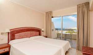 Et hotelværelse med to enkeltsenge, en træhovedgavl og natborde. En glasparti med udsigt over havet og altanmøbler. (AI-genereret tekst)