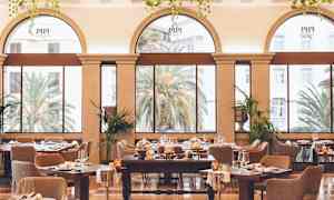 En elegant indrettet spisesal med store buede vinduer, der vender ud mod en frodig udsigt med palmer. (AI-genereret tekst)