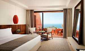 Et hotelværelse med seng, bord og siddeplads, med udsigt over havet fra en terrasse. (AI-genereret tekst)