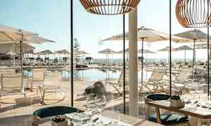 Udsigt fra en restaurant med borde og glasopdækning mod en terrasse med liggestole og parasoller ved en pool. (AI-genereret tekst)