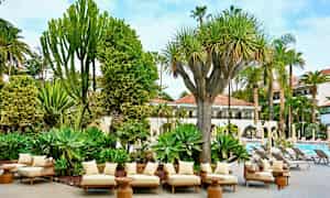 En terrasse med komfortable siddepladser omgivet af tropiske planter og udsigt over en pool. (AI-genereret tekst)