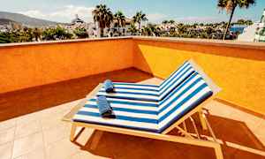 Udsigt fra en solterrasse med to solsenge med stribede blå og hvide puder samt håndklæder på, omgivet af palmer og bygninger. (AI-genereret tekst)